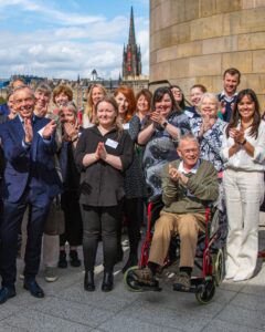 A group of MGS Business Support Programme participants smile and clap as they pose together on a balcony. Behind the group is the skyline of Edinburgh's Old Town.