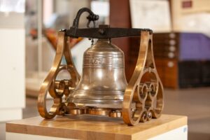 A metal bell with the numbers "1837" embossed on the side. The bell is suspended on a black bar which is held up by decorative metal supports.