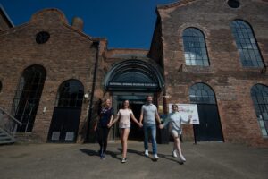 Four people with light skin - two adults, a teenager, and a child - hold hands and walk together out of a large red brick building. Above the door is the text "National Mining Museum Scotland".