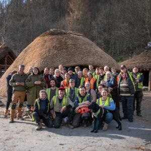 A large group of people wearing hi-vis vests are standing and sitting in front of a replica crannog and smiling at the camera.