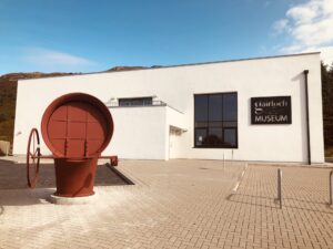 A white rectangular building with a black sign on it "Gairloch Museum". In front of it is a red metal cylinder with a wheel attached to it.