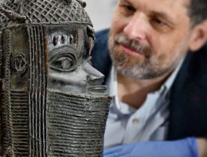 A Benin Bronze sculpture is pictured in the foreground. The sculpture depicts the head of an Oba of Benin. A person with light skin tone and dark hair is in the background observing the sculpture.