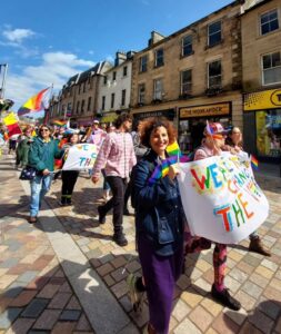 A crowd of people smile and wave rainbow Pride flags as they march down a high street on a sunny day.