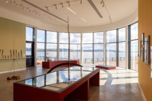 A large gallery space with a curved wall. The curved wall has floor-to-ceiling windows which offer a view of a large body of water.