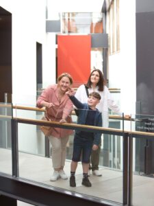 An adult with light skin and tied-back wavy red hair, an adult with light skin and medium length dark brown hair, and a child with light skin and short brown hair stand together on a raised walkway within a spacious gallery. The child is pointing to something high above them.