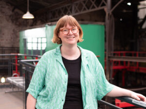 An adult with light skin, medium-length auburn hair, brown glasses, and a light green shirt.