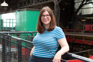 An adult with light skin, straight brown shoulder-length hair, glasses. and a blue and white striped t-shirt.