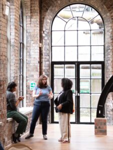 Three adults engage in conversation in a large room with tall windows and red brick walls.