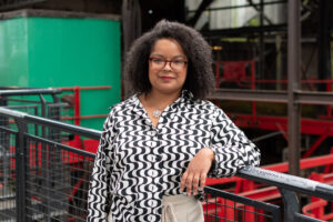 An adult with medium-tone skin, curly black shoulder-length hair, glasses, and a black and white wave-pattern shirt.