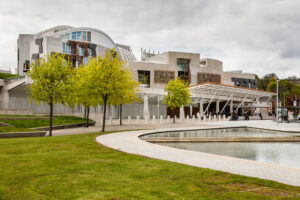 A modern concrete building with a wide awning. Outside the building are trees, a lawn, and shallow pools of water.