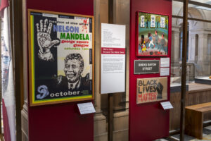 A museum display featuring a poster of South African anti-apartheid activist Nelson Mandela, a placard with an image of a raised fist and the text "Black Lives Matter", and a black sign with the text "Sheku Bayoh Street".