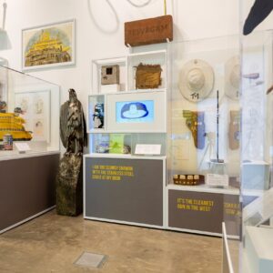 A line of display cases containing sculptures and artefacts relating to Scottish artist George Wyllie. This includes a model of a ferry, a locomotive, and a large metal sculpture of a bird of prey.