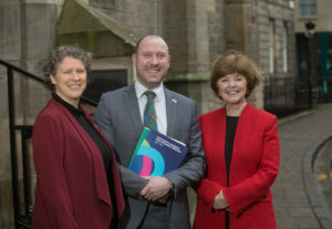 Three adults standing together in a line and smiling. The adult in the centre is holding a copy of Scotland's Museums and Galleries Strategy.