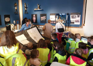 An adult with light skin and medium-length brown hair shows a map to a group of children.