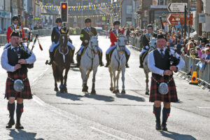 Two bagpipers and five adults on horseback parade down a street.