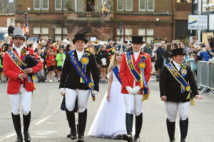 A bagpiper and three adults, all wearing traditional riding uniforms with blue sashes and ribbons, walk alongside a child who is wearing a long white dress and a crown.