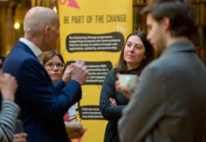 A group of four adults stand and engage in conversation in front of a yellow banner with the text 'Be part of the change'.